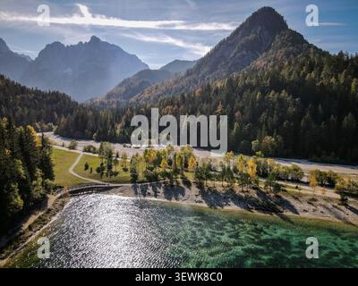 Eine Weitwinkelaufnahme fängt den Fusine-Inferiore-See im Vordergrund ein, mit einem Herbstwald und den Julischen Alpen im Hintergrund. Dieses Bild ist Suita Stockfoto