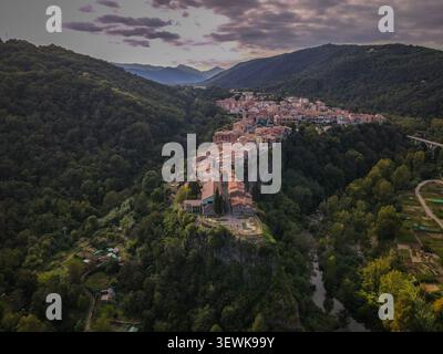 Aus der Vogelperspektive sehen Sie das Dorf Castellfollit de la Roca und die Kirche Sant Salvador auf einem Basaltkliff, umgeben von einem bewaldeten Tal in der Region Stockfoto