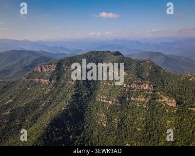 Eine Weitwinkelaufnahme aus der Luft fängt ein riesiges waldbedecktes Bergmassiv in der Region Osona, Katalonien, Spanien, unter einem klaren blauen Himmel ein. Dieses Bild ist geeignet Stockfoto