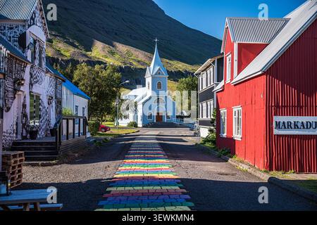 Der ikonische regenbogenfarbene Backsteinpfad führt in Richtung der berühmten blauen Holzkirche im malerischen Dorf Seydisfjordur in Ost-Island. Stockfoto