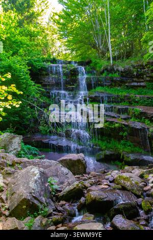 Vertikaler Blick auf einen malerischen, mehrstufigen Wasserfall, der über moosige Felsen in einem lebhaften grünen Wald fließt, wunderschöne natürliche Landschaft eines hidd Stockfoto