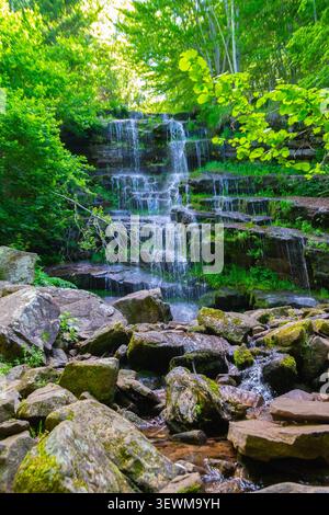 Vertikaler Blick auf einen malerischen, mehrstufigen Wasserfall, der über moosige Felsen in einem lebhaften grünen Wald fließt, wunderschöne natürliche Landschaft eines hidd Stockfoto