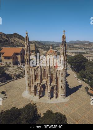 Blick aus der Vogelperspektive auf den Santuario de Santa Maria Magdalena mit seinen markanten Steintürmen und Terrakotta-Dach, umgeben von einem gepflasterten platz und einem trockenen Hügel Stockfoto