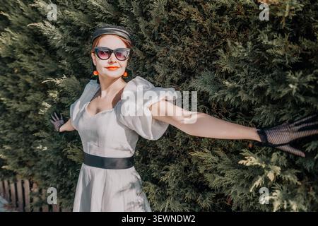 Frau weiß Kleid Mode posiert mit Armen gestreckt gegen eine grüne Gartenhecke an einem hellen sonnigen Sommertag Stockfoto