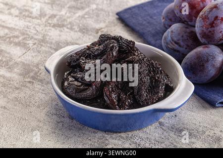 Getrocknete Pflaumen in blauer Keramikschale neben frischen Pflaumen auf strukturierter grauer Oberfläche. Natürliches Licht hebt dunkle violette, dunkle Brauntöne hervor Stockfoto