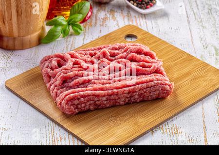 Frisches rohes Hackfleisch auf Holzschneidebrett. Hintergrund: Basilikumblätter, Pfeffer, Holzmühle. Sanfte Beleuchtung hebt rotes Fleisch hervor Stockfoto