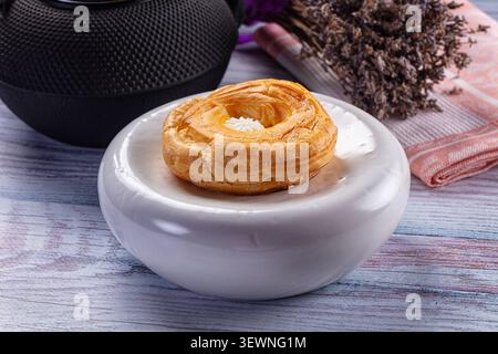 Goldener Blätterteig mit Creme-Mitte auf weißem Teller. Rustikaler Holztisch mit getrocknetem, lavendelschwarzem Lautsprecher im Hintergrund. Weiches natürliches Licht, warm Stockfoto