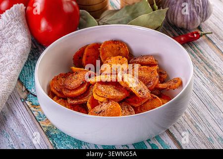 Geröstete Karottenscheiben in weißer Schüssel umgeben von roten Pfeffer Knoblauch Lorbeerblättern auf verwitterter Holzoberfläche warme Beleuchtung Erdtöne rustikales Küchenset Stockfoto