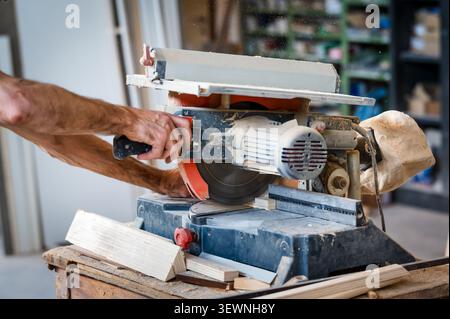 Ein Zimmermann betreibt eine Gehrungssäge, um in einer Holzwerkstatt Holzstücke zu schneiden, wobei Sägespäne fliegen und Holzreste auf der Werkbank sichtbar sind. Stockfoto