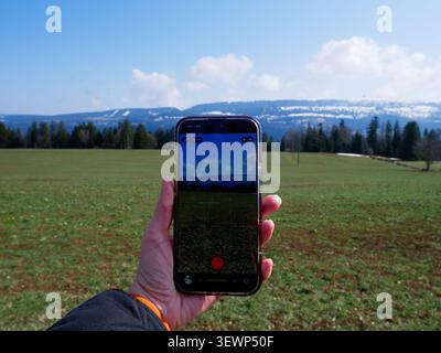 Die Person mit einem Smartphone in der Hand erfasst eine malerische Landschaft mit Bergen und einem grünen Feld, mit einem hellblauen Himmel und verstreuten Wolken Stockfoto