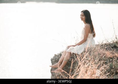 Asiatische Frau in weißem Spitzenkleid sitzt auf felsigem Seeufer in sanftem Sonnenlicht, genießt ruhige Natur, ruhige Reflexion, friedliche Einsamkeit, Entspannung, wir Stockfoto