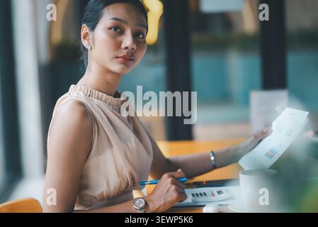 Selbstbewusste asiatische Geschäftsfrau mit Finanzbericht und Stift am Café-Schreibtisch mit Taschenrechner und Kaffee, repräsentiert Buchhaltungsarbeit, Datenüberprüfung, Kumpel Stockfoto