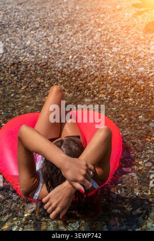 Strand schwimmende Sommerfrau, die sich auf rosa aufblasbarem Ring im klaren Meerwasser in der Nähe eines Kieselufers unter hellem Sonnenlicht entspannt Stockfoto