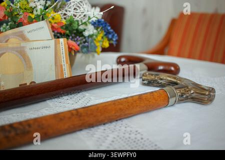 Eine Nahaufnahme von zwei traditionellen hölzernen Gehstöcken, die auf einer weißen Tischdecke neben fünfzig Euro-Banknoten und einem bunten Blumenstrauß liegen. A Konz Stockfoto