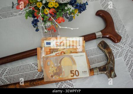 Ein Hochwinkel von 50 Euro-Scheinen, die über zwei Wanderstöcken auf einer weißen Tischdecke mit Spitze neben Blumen platziert wurden, symbolisiert die finanziellen Kosten von Stockfoto