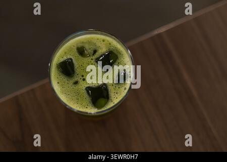 Scharfer, detaillierter Blick auf ein erfrischendes grünes Matcha-Teegetränk, serviert mit Eiswürfeln und dichtem Schaum in einem klaren Glas auf einem dunklen Holztisch. Stockfoto