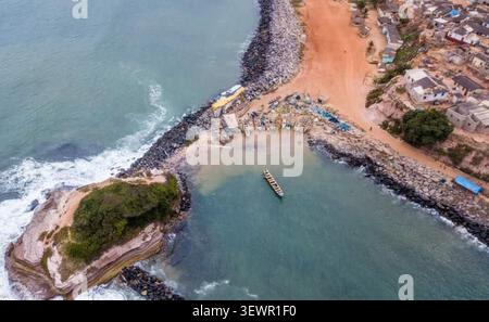 Aus der Vogelperspektive auf eine Küstenmole mit kleinen Fischerbooten, die in einer geschützten Bucht vor Anker liegen, neben einem kleinen Dorf mit roten Feldwegen in Ghana. Stockfoto