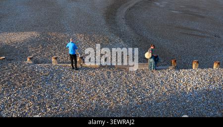 Mann mit blauem Trainingsanzug und schwarzer Hose, der allein bei Sonnenuntergang an einem Kieselstrand steht, während eine Frau in langem Rock in der Nähe mit dem Blick weg sitzt Stockfoto