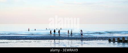 Badegäste am Strand und im Meer bei Sonnenuntergang im Juni mit gedämpften Farben am Himmel dahinter Stockfoto
