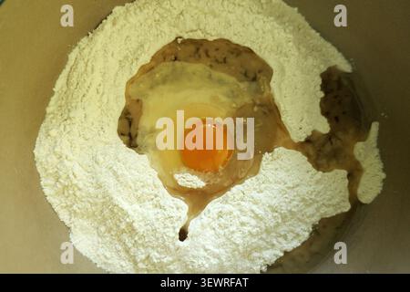 Mehl, rohes Ei und flüssige Zutaten werden in einer Schüssel gemischt, um die wesentlichen Schritte der Teigzubereitung zum Backen oder Kochen darzustellen Stockfoto