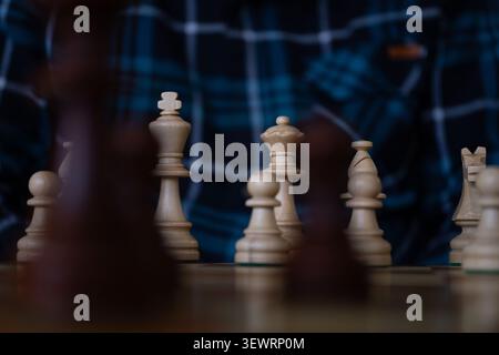 Hölzerne Schachfiguren auf einem Schachbrett mit einem verschwommenen Spieler im Hintergrund, die strategische Planung, Führung, Analyse und Dekisi symbolisieren Stockfoto