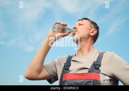 Mann im Overall, der während der Arbeitspause mit blauem Himmel hydriert Stockfoto