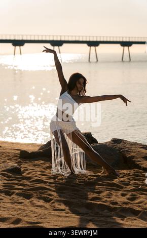 Eine elegante junge kubanische Frau, hinterleuchtet, tanzt bei Sonnenaufgang am Strand El Coco in Badalona, trägt ein weißes Top und einen Fransen-Makramee-Rock, mit dem Pont del Stockfoto
