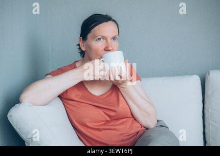 Nachdenkliche Frau mittleren Alters trinkt Kaffee auf dem Balkonsofa während der morgendlichen Routine und genießt einen ruhigen Moment der Reflexion und Entspannung. Selektiver Fokus Stockfoto