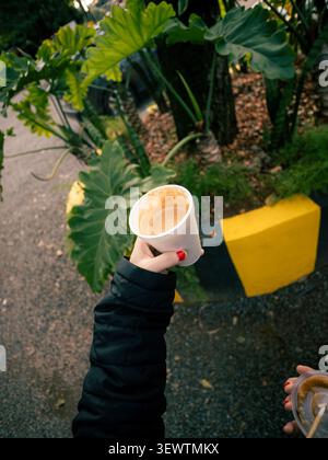 Die Hand hält eine weiße Kaffeetasse mit rotem nagellack. Eine andere Hand greift eine Schüssel in der Nähe. Große grüne Blätter füllen den Hintergrund. Gelbe Bordsteinkante markiert den p Stockfoto