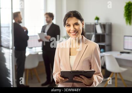 Lächelnde Geschäftsfrau mit Klemmbrett im modernen Büro, selbstbewusstes Porträt Stockfoto