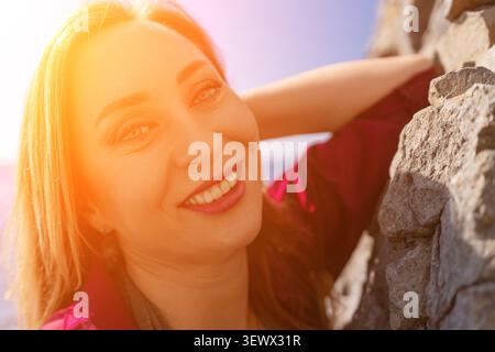Frauenporträt Lächeln schöne Erwachsene Frau, die glücklich lächelt, während sie sich unter hellem Sonnenlicht am Strand gegen eine Steinmauer lehnt Stockfoto