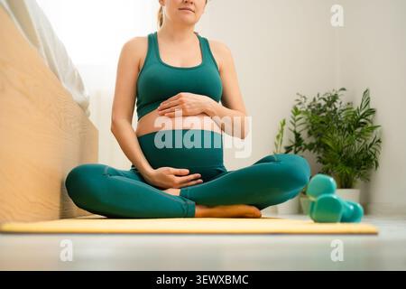 Schwangere Frau sitzt auf Yogamatte in friedlicher pränataler Yogaposition zu Hause. Erwartende Mutter, die Hand auf den Bauch legt, in achtsamer Meditationsstellung. Mutter-zu-werdende übt ruhiges Yoga zur Geburtsvorbereitung Stockfoto