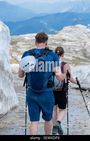 Nahaufnahme von zwei Wanderern mit Rucksäcken und Trekkingstöcken, die an einer hohen Schneemauer vorbeilaufen, und ein Paar, das ein aktives alpines Abenteuer im Sommer genießt Stockfoto