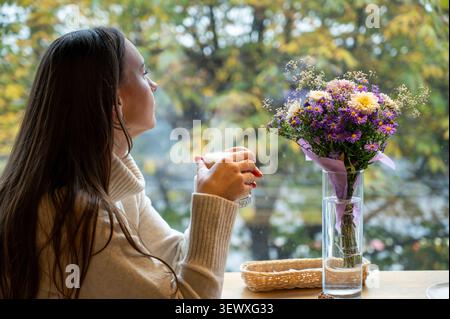 Traumhafte Dame mit einer Tasse Kaffee am Holztisch mit Blumenstrauß und Desserts im Restaurant gemütliche Herbststadt-Atmosphäre im Hintergrund Stockfoto
