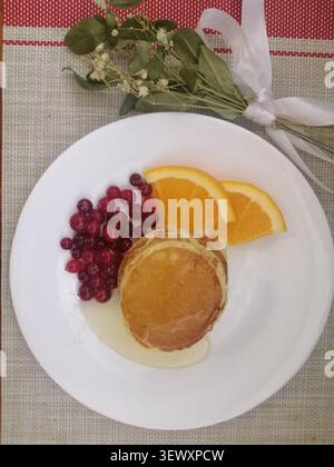 Ein Blick von oben auf einen Stapel flauschiger Pfannkuchen, serviert auf einem weißen Teller mit frischen roten Preiselbeeren, saftigen Orangenscheiben und Honig darauf Stockfoto