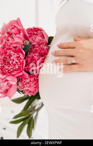 Ein zartes Porträt, das eine schwangere Frau in einem weißen Kleid fängt, die sanft ihren Bauch neben einem leuchtenden Blumenstrauß rosafarbener Pfingstrosen hält. Stockfoto