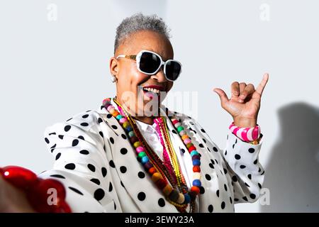 Schwarze Seniorin trägt moderne Kleidung Porträt auf Hintergrund - stilvolle Seniorin lächelt fröhlich. Sonnenbrille tragen. Bunte Perlen. Und A Stockfoto