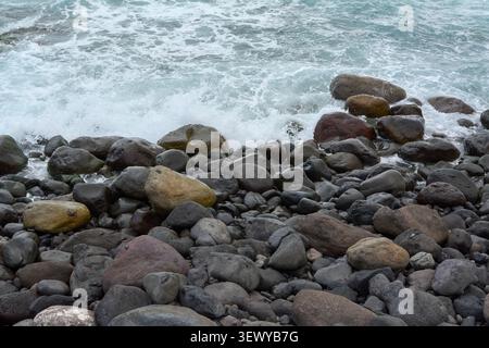 Ozeanwellen treffen auf eine felsige Küste, die mit großen, abgerundeten Vulkansteinen und Kieselsteinen bedeckt ist, in Agaete, Spanien. Stockfoto