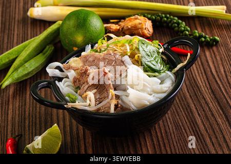 Nahaufnahme mit traditioneller vietnamesischer Rindfleischnudelsuppe. Schwarze Schüssel auf dunklem Holztisch mit frischen Zutaten wie Limette, Chili und Kräutern. Appetitlich Stockfoto