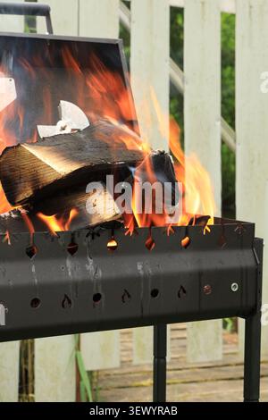 Ein Fragment eines Grills mit einem Haufen Brennholz, das mit einer orangen Flamme vor dem Hintergrund eines weißen Zauns brennt Stockfoto