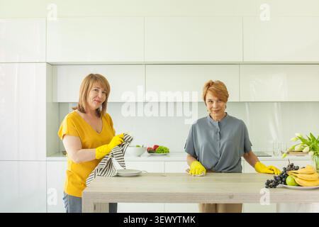 Zwei Frauen putzen die Küche und bereiten tagsüber Essen zu. Sie tragen gelbe Handschuhe, während sie den Tisch in einem hellen Raum mit Essen organisieren Stockfoto