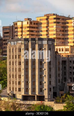 Ein hohes, verwittertes Betongebäude steht im Vordergrund. Dahinter erheben sich größere, orange-braune Wohnblöcke. Sonnenlicht hebt das Alter des Gebäudes hervor Stockfoto