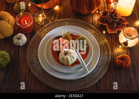 Herbstlicher Tisch, der eine gemütliche Atmosphäre für ein Weihnachtsessen schafft, mit Teller, goldenem Besteck, Stoffkürbissen, Glühkerzen, und Zeichenfolge Stockfoto