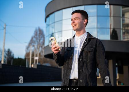 Junger Mann, der ein Smartphone vor einem modernen Bürogebäude hält, während er eine mobile Banking-Anwendung verwendet. Mobile Banking- und Fintech-Services für immer Stockfoto