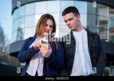 Junge Geschäftskollegen, die während des Arbeitstages gemeinsam ein Smartphone außerhalb eines modernen Bürogebäudes verwenden. Teamarbeit und professionelle Zusammenarbeit für Stockfoto