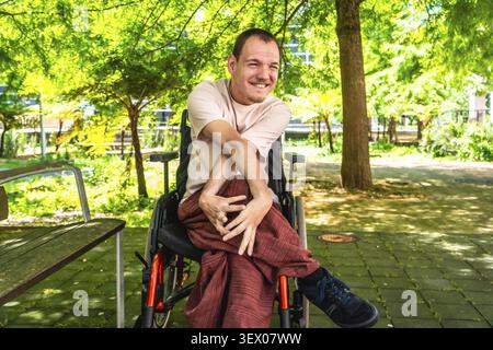Lächelnder Mann mit Zerebralparese, der im Rollstuhl sitzt, sich glücklich fühlt und tagsüber eine zugängliche Outdoor-Park-Umgebung genießt Stockfoto