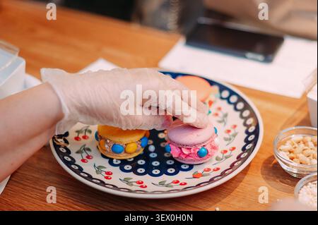 Hand in Schutzhandschuh zur Dekoration bunter gefüllter Macarons auf Keramikplatte in der Konditorei Stockfoto
