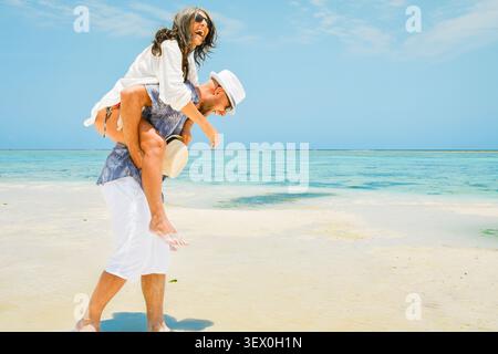 Multi-ethnisches glückliches Paar, das Spaß beim Huckepack-Reiten am weißen Sandstrand hat. Kaukasischer Mann und Frau mittleren Alters im spielerischen Moment am Ufer bei Summe Stockfoto