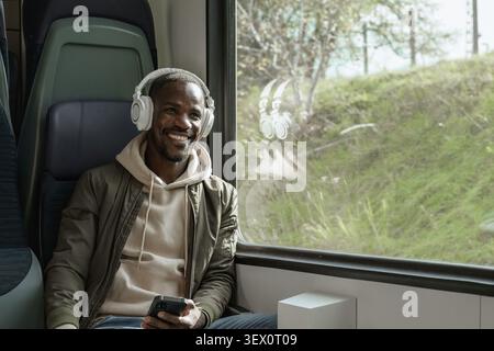Ein afroamerikanischer Mann reist mit dem Zug, lächelt und verwendet sein Smartphone und Kopfhörer, während er während seiner Reise Musik hört. Stockfoto