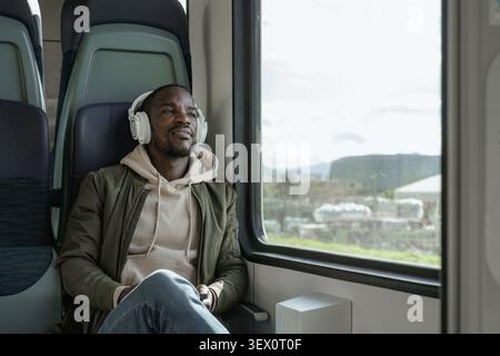 Ein afroamerikanischer Mann reist mit dem Zug, lächelt, während er seine Kopfhörer benutzt und während seiner Reise Musik hört. Stockfoto